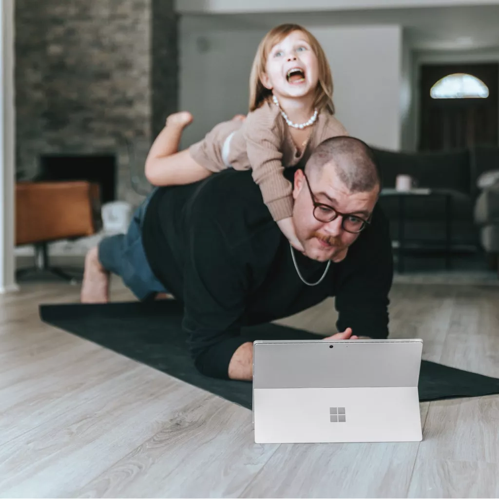 A father trying to do yoga, with his daughter having fun on his back.