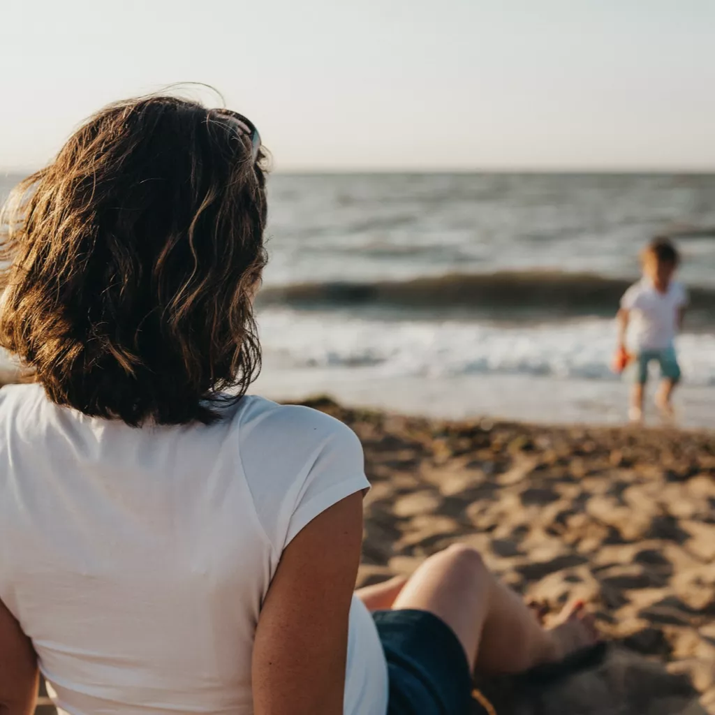 A lady sat on a beach watching her child play.